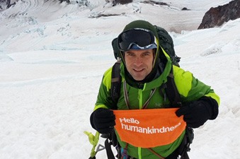 Michael Seagraves poses on a snowy mountain trail  with a Hello Humankindness banner