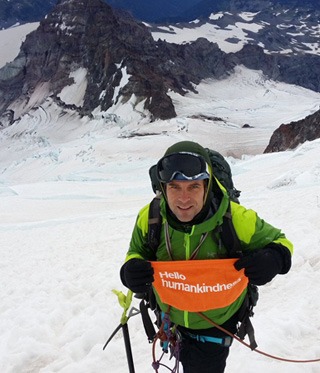 Michael Seagraves poses on a snowy mountain trail  with a Hello Humankindness banner