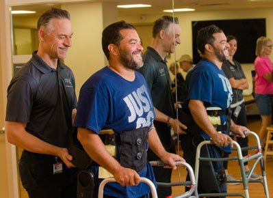A therapist help guide a smiling man in a back brace and walker