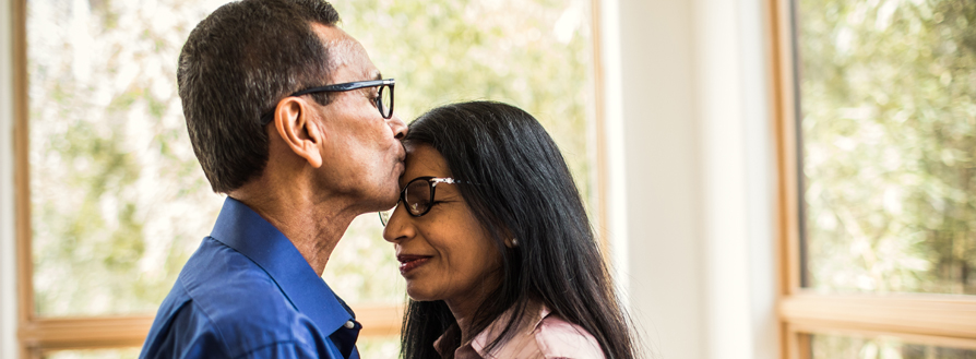 One spouse kissing the other on the forehead in front of windows looking upon trees.
